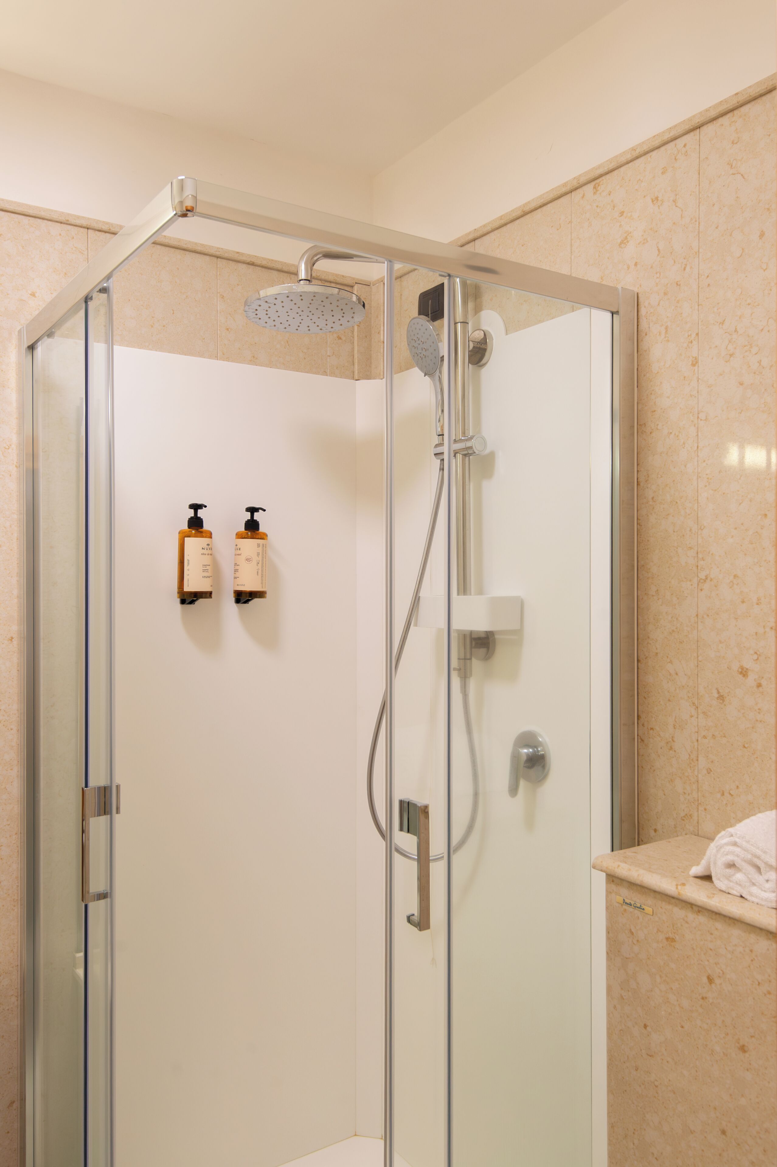 Ducha con cerramiento de vidrio, cabezal de ducha manual, secador de manos en la pared y dos botellas de champú.