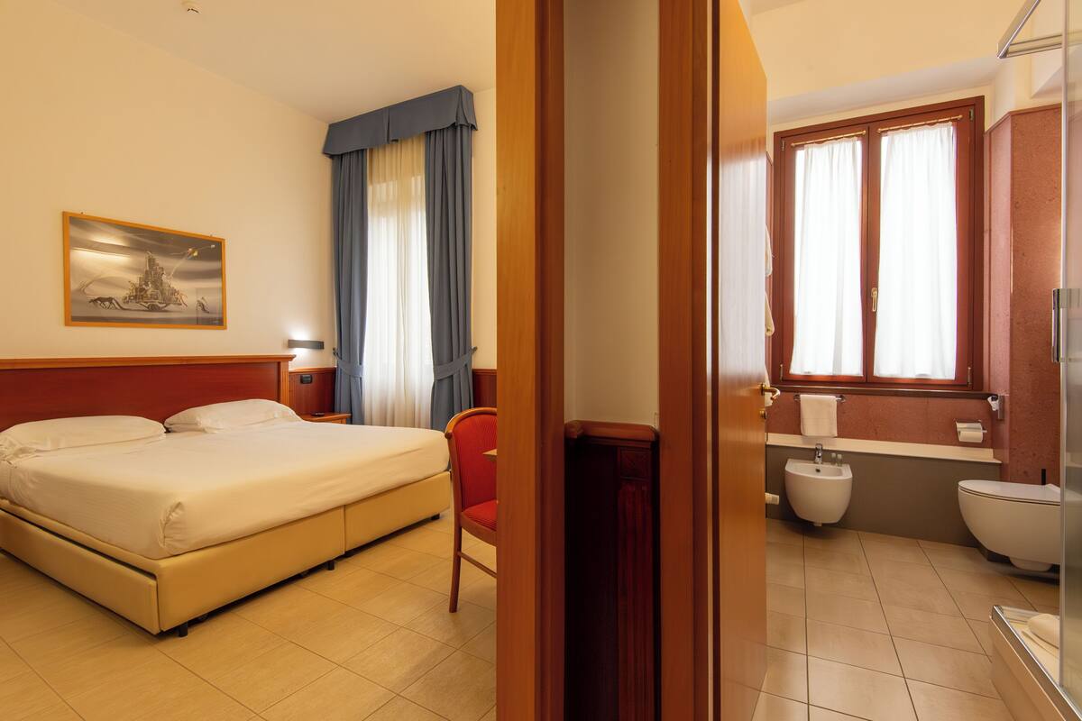 Une chambre d’hôtel comprenant un lit, une chaise, une salle de bain avec un bidet et une fenêtre avec des rideaux.