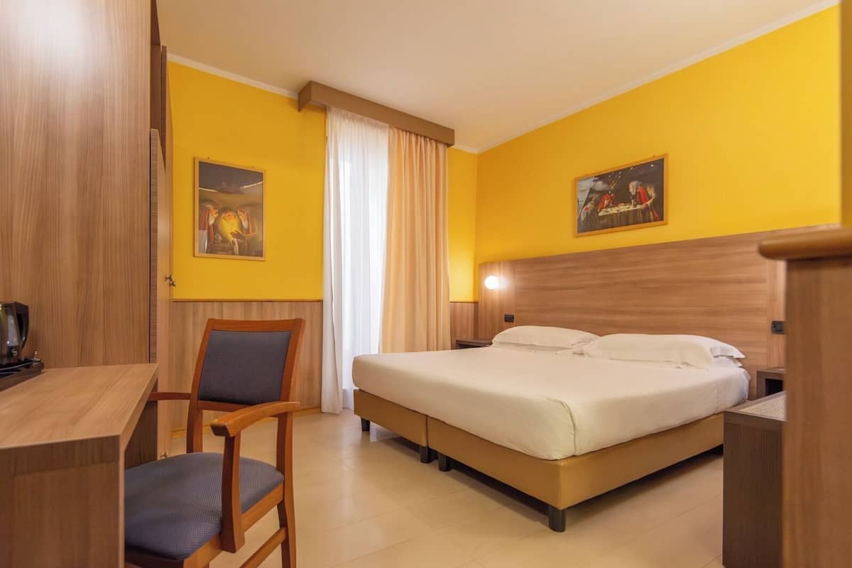 Une chambre d’hôtel avec un lit, un bureau, une chaise, une table de chevet et un tableau au mur.