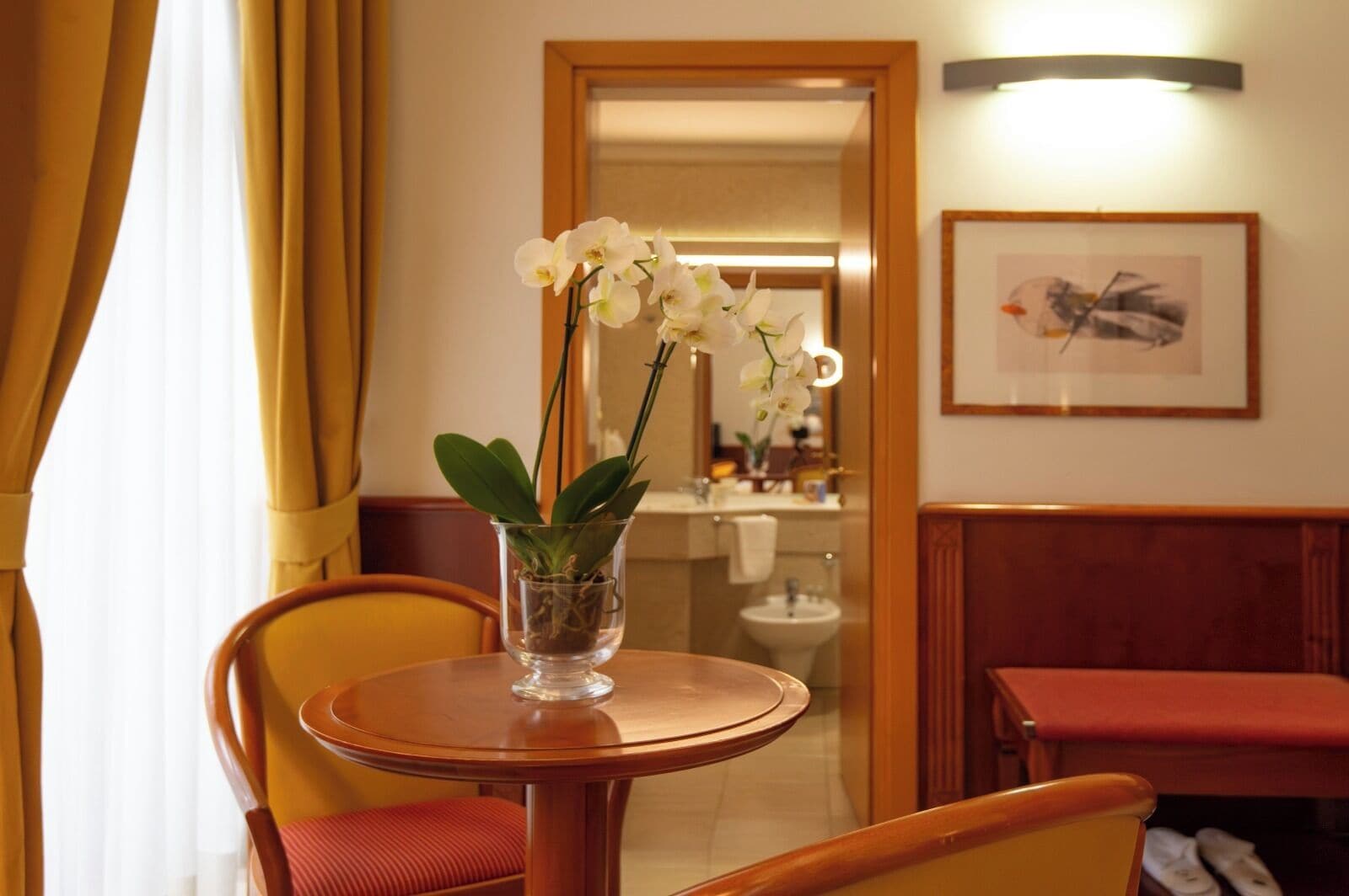 A hotel room with a round wooden table, a chair, a glass vase with white orchids, a framed picture on the wall, and a bathroom visible through an open door.