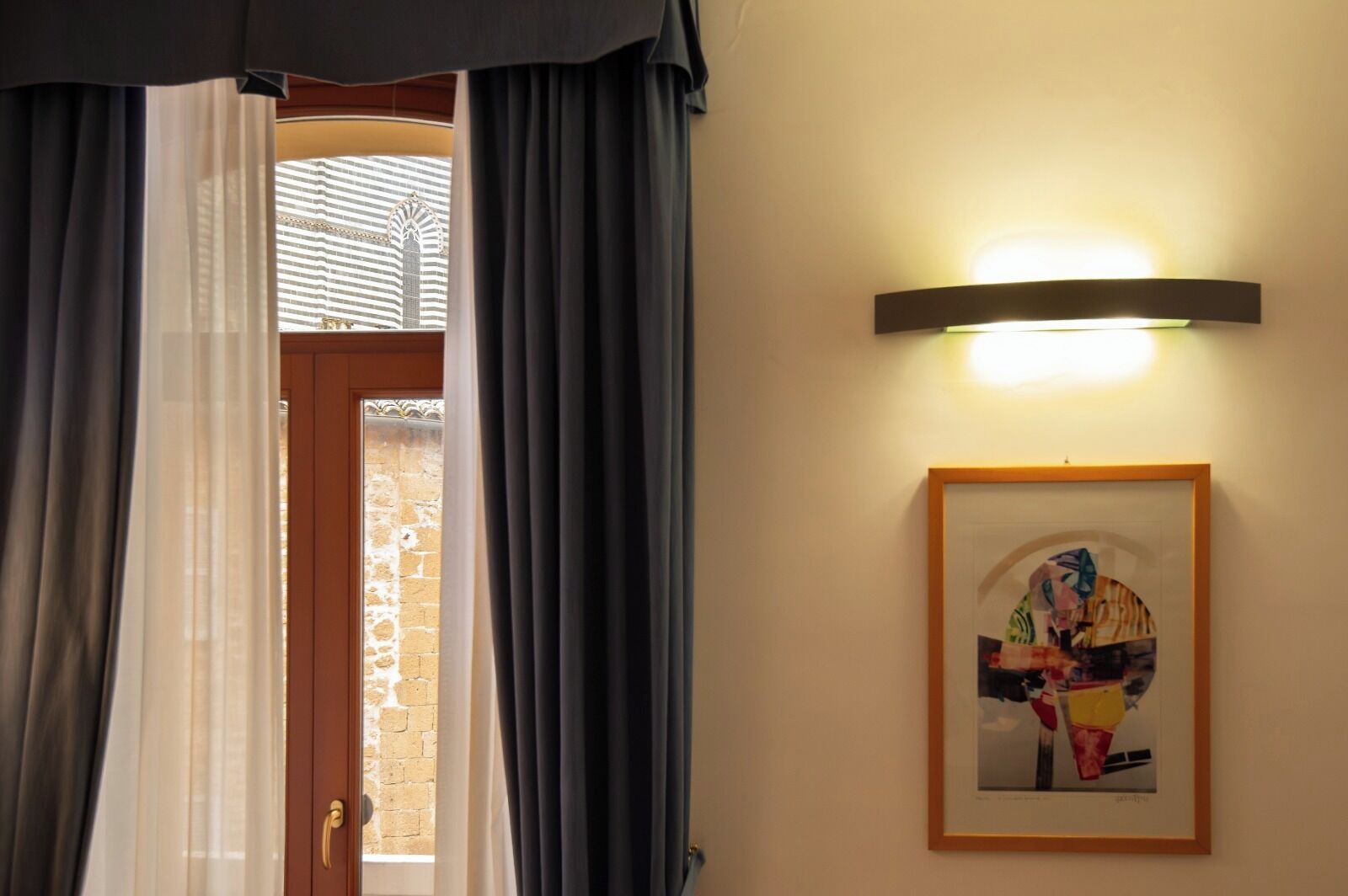 Una camera d'albergo con una porta, tende, un quadro incorniciato e un punto luce a parete.