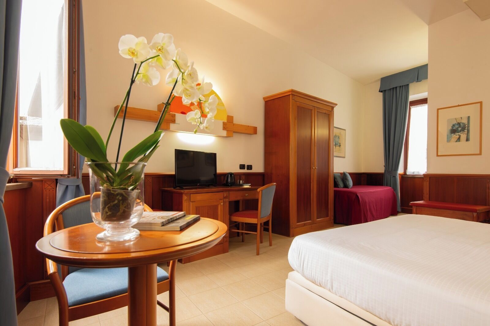A hotel room with a bed, desk, chair, television, wardrobe, and a potted orchid.