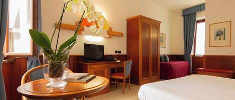 Habitación de hotel con cama, escritorio, silla, televisor, armario y una orquídea en maceta.