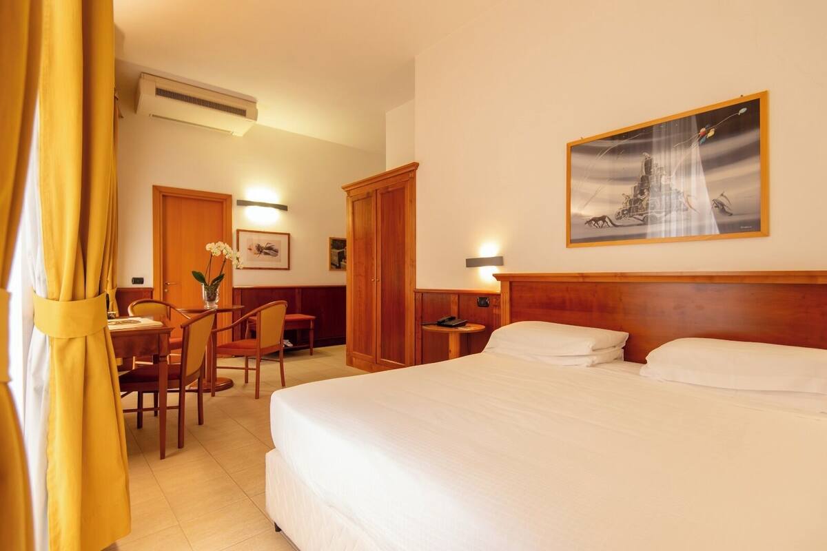 Une chambre d’hôtel avec un lit, un bureau et une chaise, une petite table avec un vase, et un tableau au mur.