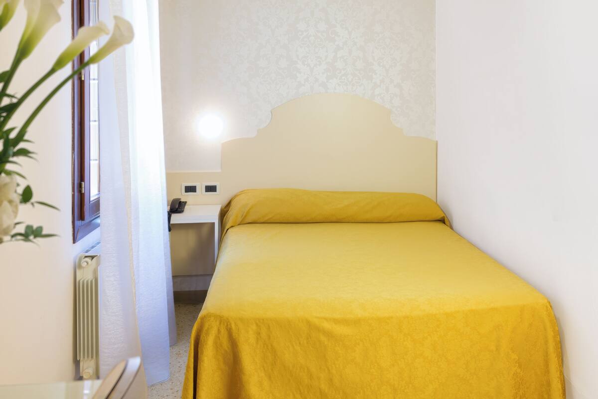 A small, neatly made bed with a yellow bedspread in a room with white walls and a patterned wallpaper.