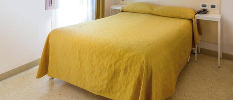 A neatly made bed with a yellow bedspread in a room with a speckled floor, a bedside table with a phone, and a wall-mounted light fixture.