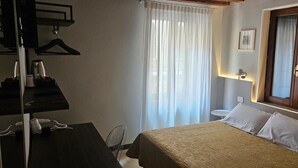 A hotel room with a bed, a desk with a telephone, a chair, a window with curtains, and a wall-mounted lamp. - Hotel Adriatico (Venice)