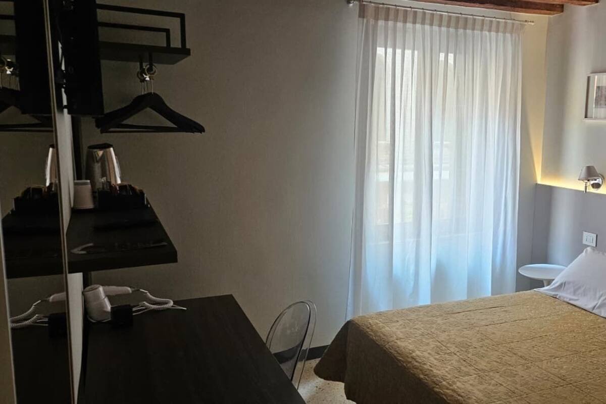 A hotel room with a bed, a desk with a telephone, a chair, a window with curtains, and a wall-mounted lamp.