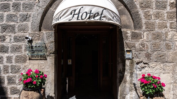 A hotel entrance with a stone facade, an arched doorway, and two flower pots with pink flowers. - Hotel Corso (Orvieto)