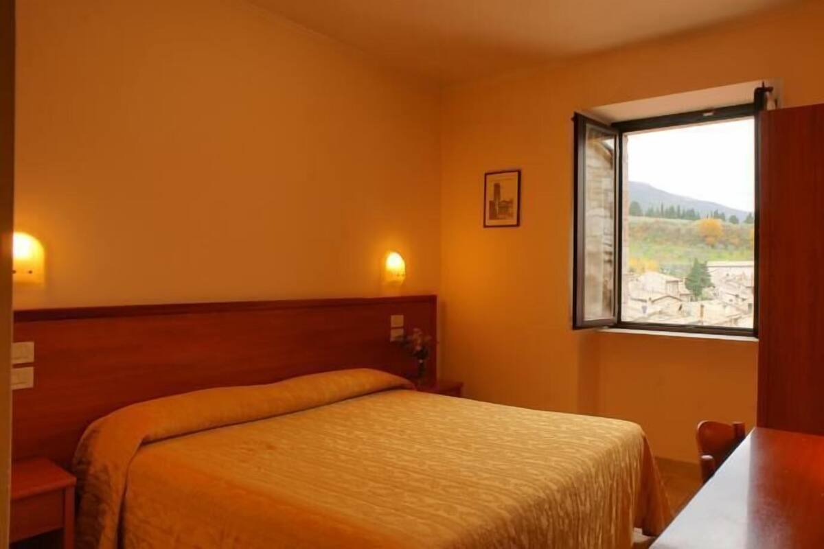 A hotel room with a bed, a desk, a window with a view of a landscape, and wall-mounted lights.