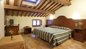Un dormitorio con techo de madera, una cama con colcha estampada, una mesita de noche, una pequeña mesa con un jarrón de flores y una ventana con cortinas.