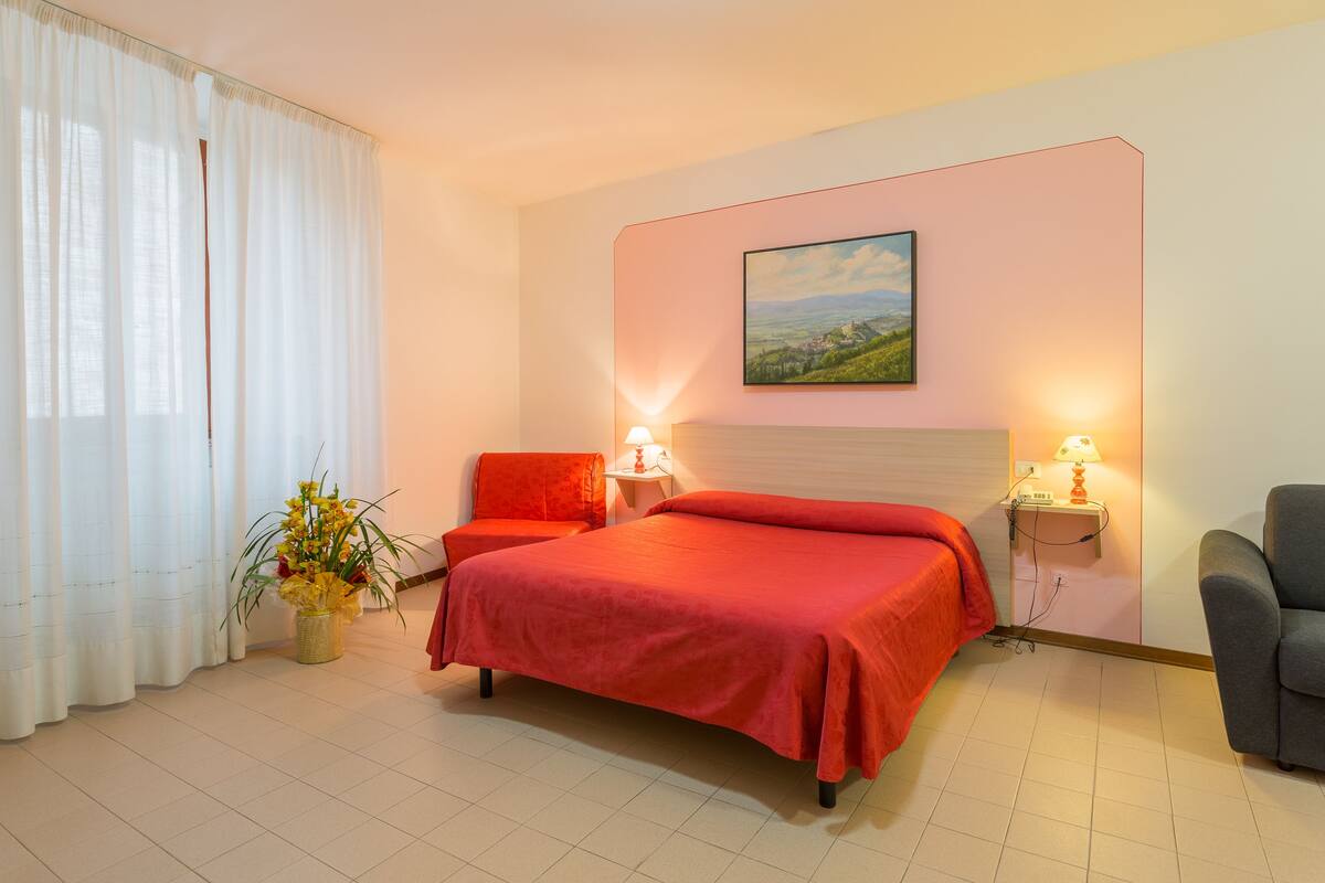 A hotel room with a bed, two bedside tables with lamps, a chair, a potted plant, and a painting on the wall.