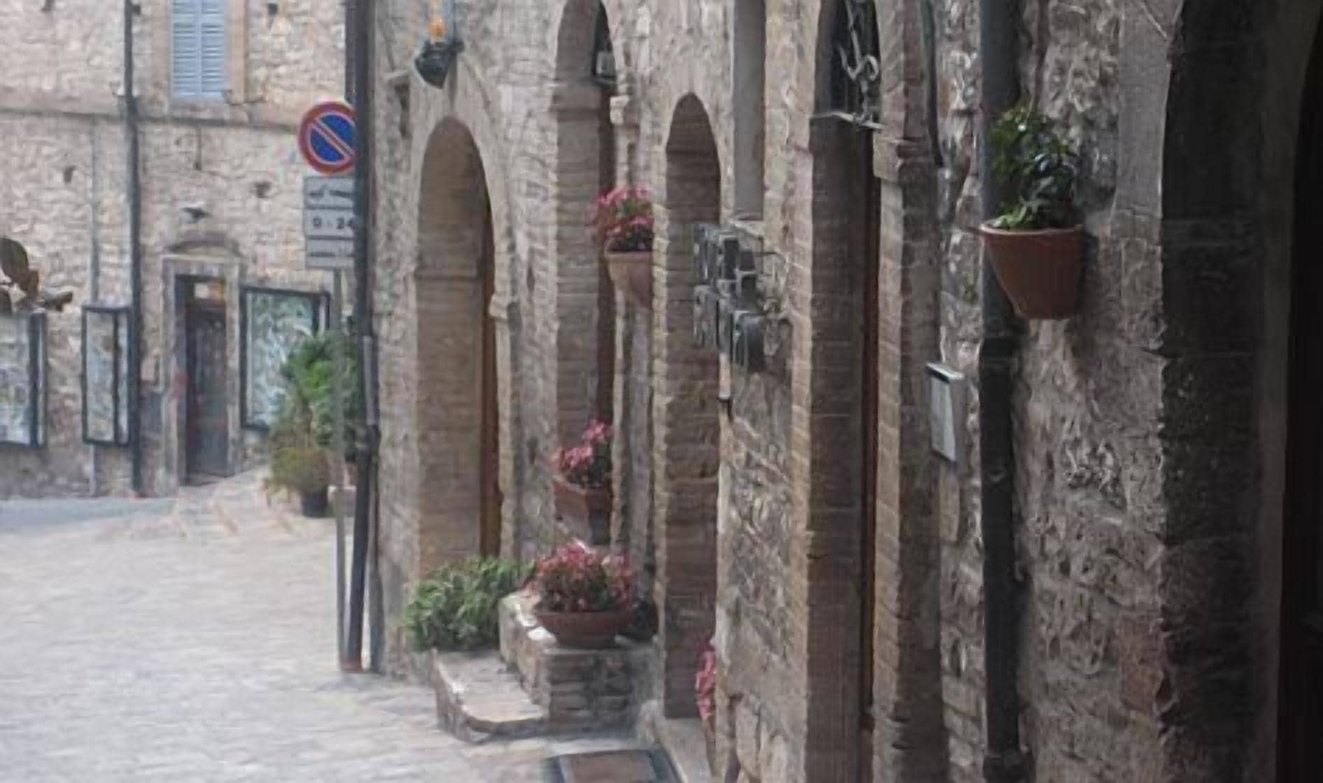 Una stretta strada acciottolata con edifici in pietra, portali ad arco e fioriere pensili.
