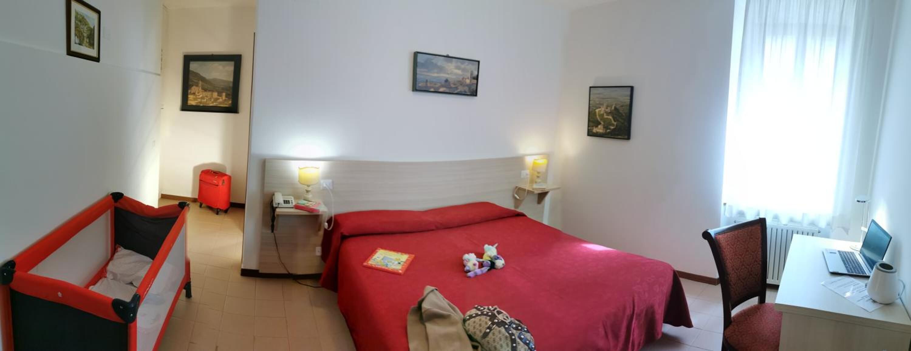 Een hotelkamer met een bed, een bureau met een laptop, een stoel, een klein tafeltje met een lamp en een rode koffer.