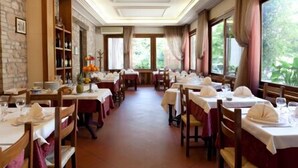 A restaurant with long tables set for dining, a brick wall on one side, and large windows on the other. - Porziuncola (Assisi)
