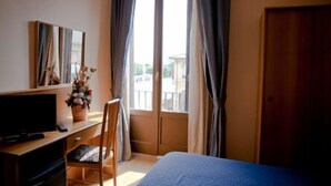A hotel room with a bed, a desk with a computer, a chair, a window with curtains, and a door. - Porziuncola (Assisi)
