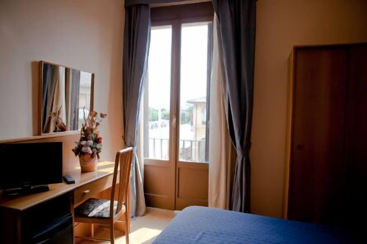 Ein Hotelzimmer mit Bett, Schreibtisch mit Computer, Stuhl, Fenster mit Vorhängen und einer Tür.