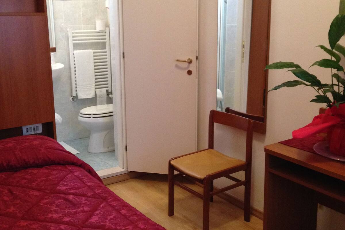 A bedroom with a bed, a wooden chair, a desk, a mirror, and a door leading to a bathroom.
