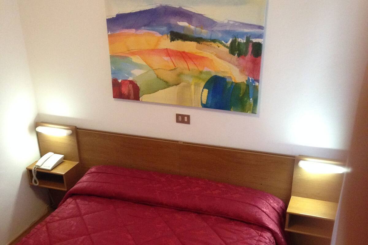 A hotel room with a wooden bed, red bedding, two bedside lamps, a nightstand with a phone, and a painting on the wall.