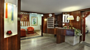 A reception area with a wooden reception desk, a clock, a framed floral artwork, and a red armchair. - Hotel Caprera (Venice)