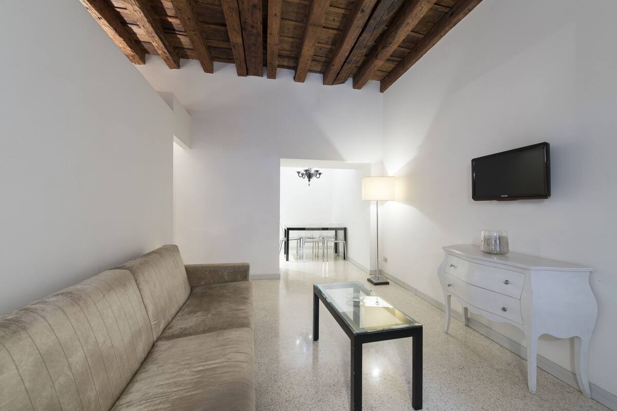 Una sala moderna con un sofá, una mesa de centro de vidrio, un aparador blanco y un televisor de pantalla plana montado en la pared.
