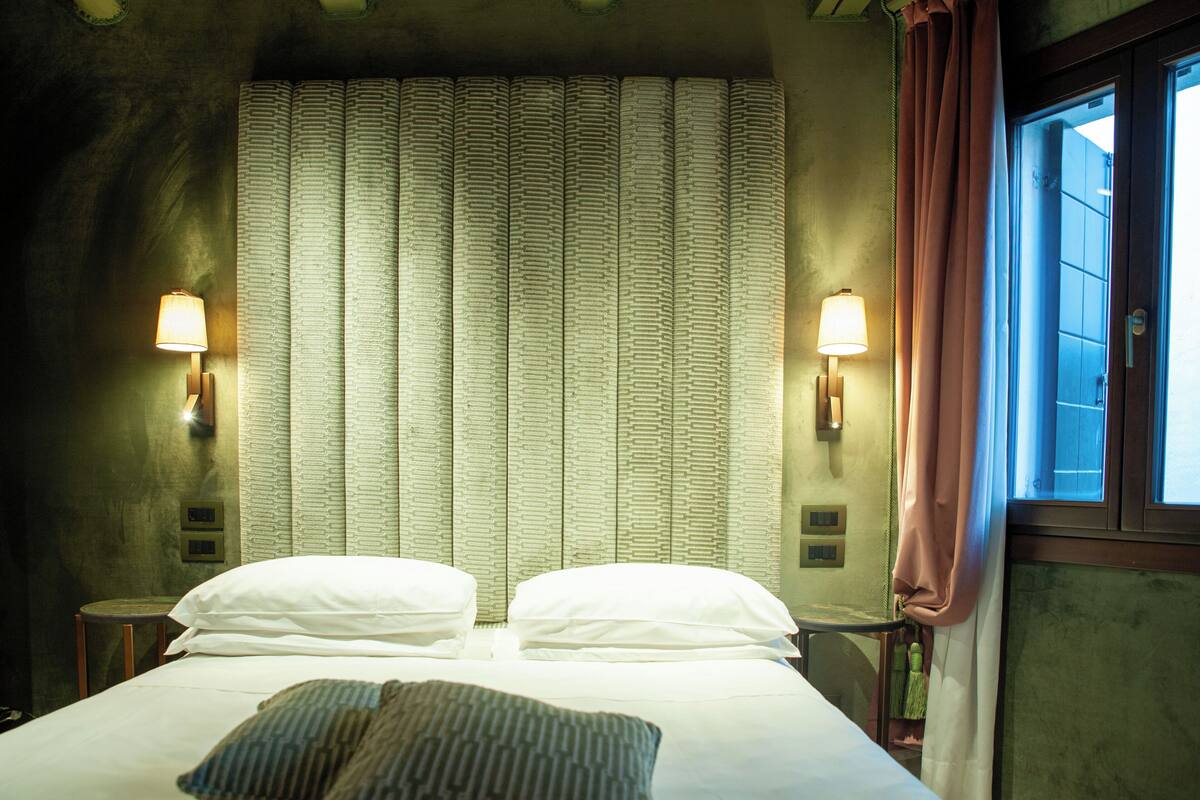 Una habitación de hotel con una cama, dos lámparas de noche, un ventanal con cortinas y un cortinaje estampado.
