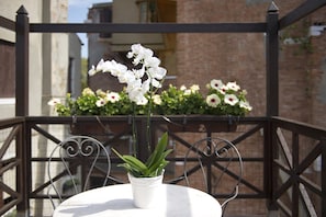 Una orquídea blanca en maceta sobre una mesa redonda con una silla metálica, frente a una pared de ladrillo y una barandilla de metal.
