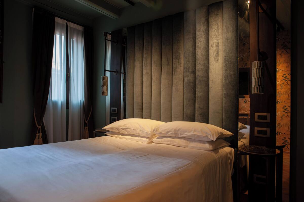 Una habitación de hotel con cama, cortinas, una lámpara fijada a la pared y una mesita.