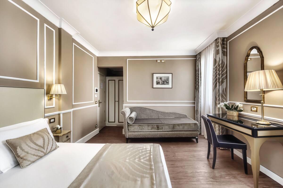 Ein Hotelzimmer mit einem Bett, einem Schreibtisch, einem Stuhl, einer Schreibtischlampe, einem gerahmten Bild an der Wand und einer Tür zu einem anderen Raum.