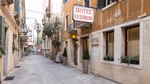A narrow street with a hotel named "HOTEL GUERRINI" on the right. There are potted plants along the sidewalk and a clear view of the street. - Hotel Guerrini (Venice)