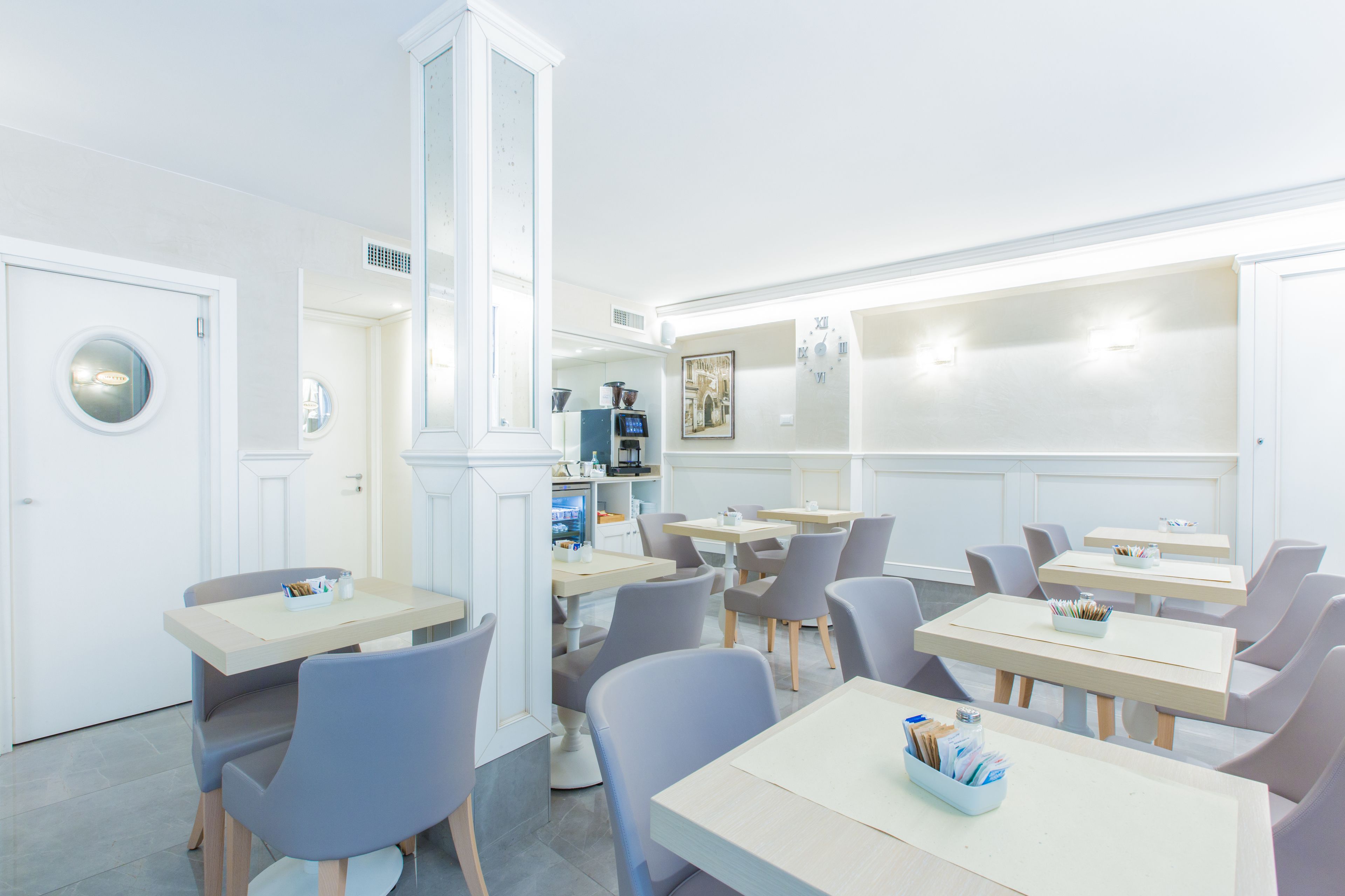 A modern restaurant with white walls, grey chairs, and wooden tables. There are multiple tables and chairs arranged neatly.