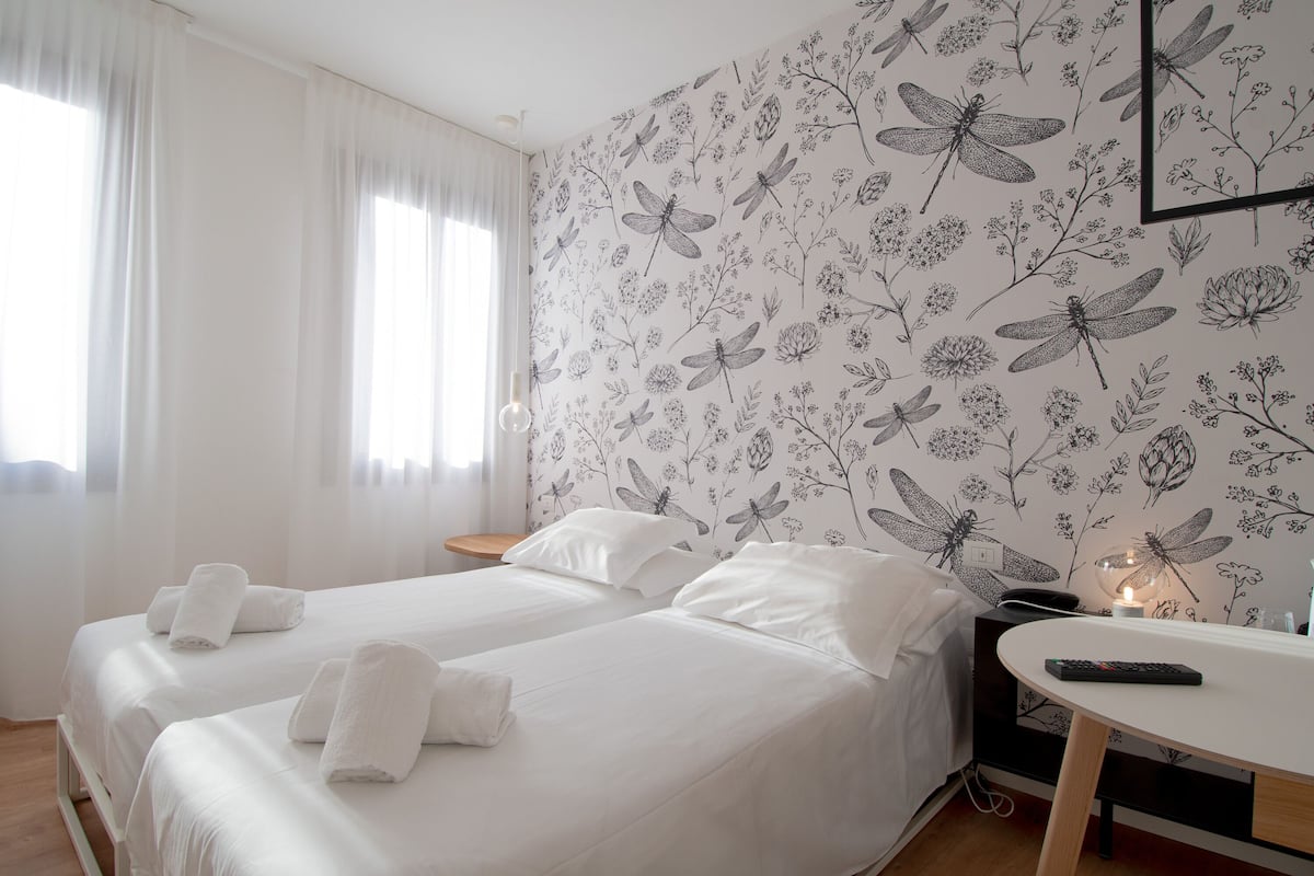 A hotel room with two beds, white linens, a desk with a remote control, and a wall with a floral and dragonfly pattern.