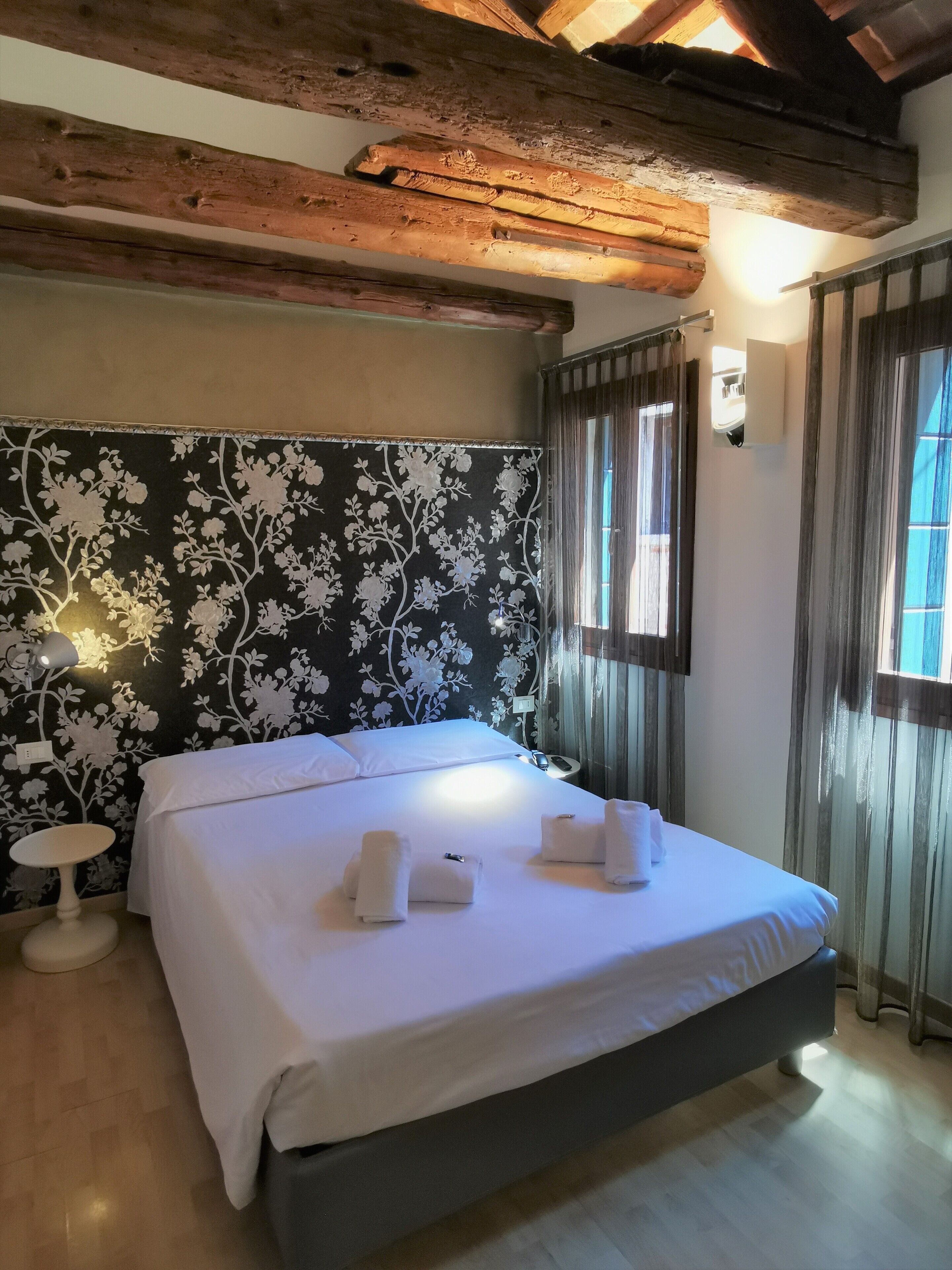A bedroom with a bed, white towels, a small bedside table, and a wall with a floral pattern.