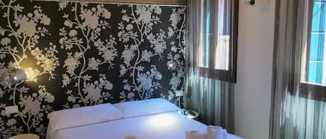 A bedroom with a bed, white towels, a small bedside table, and a wall with a floral pattern.