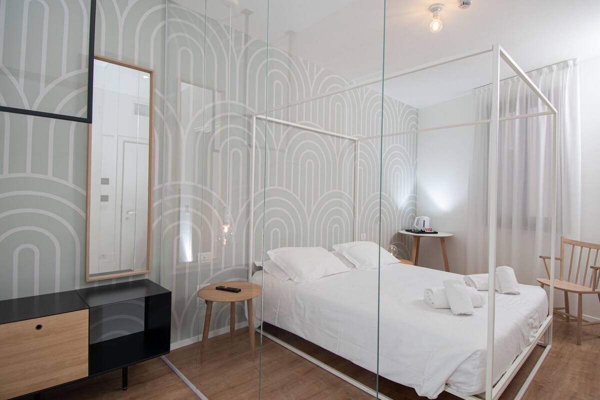 A modern bedroom with a glass partition bed, a wooden bedside table, a small round table, and a chair.