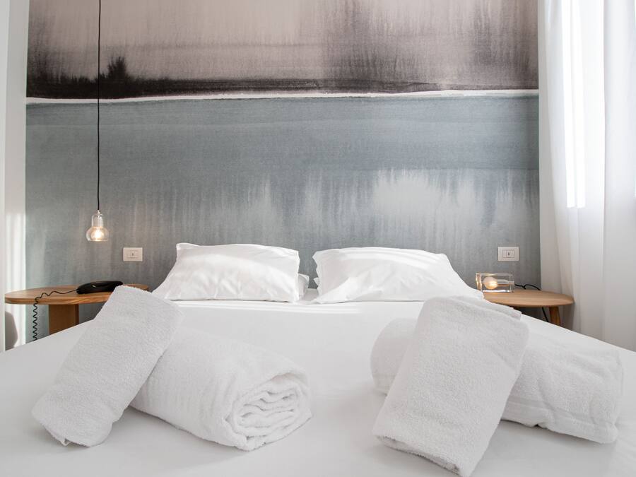Una cama bien hecha con ropa de cama y toallas blancas, una mesita de noche con una lámpara y un cuadro en la pared.