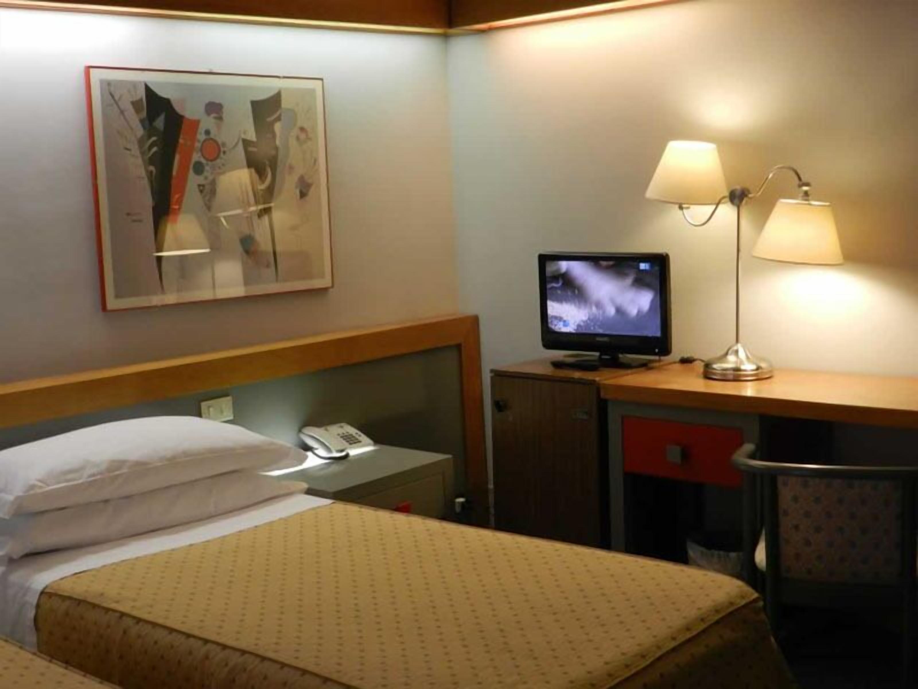 Ett hotellrum med två sängar, ett skrivbord med en dator och en telefon, en lampa och en tavla på väggen.