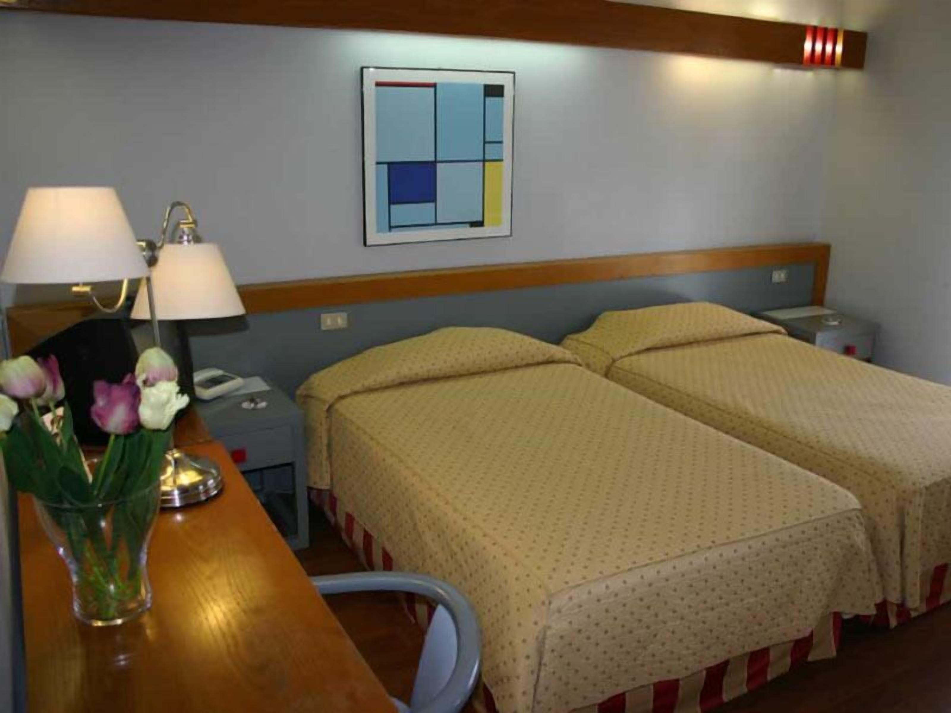Habitación de hotel con dos camas, una mesita de noche, una lámpara y un florero con flores.