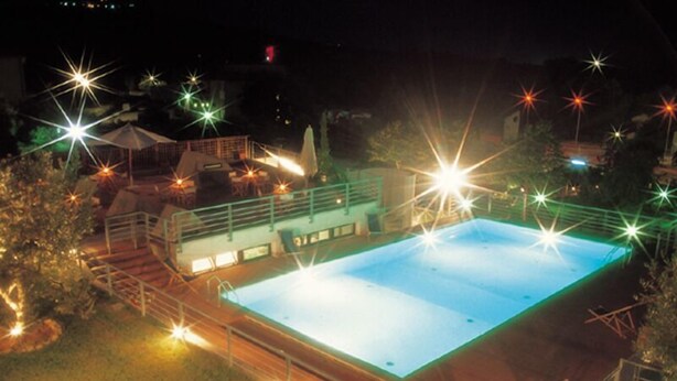 A rectangular swimming pool with a surrounding deck, surrounded by trees and a building with a green roof. - Umbria (Attigliano)