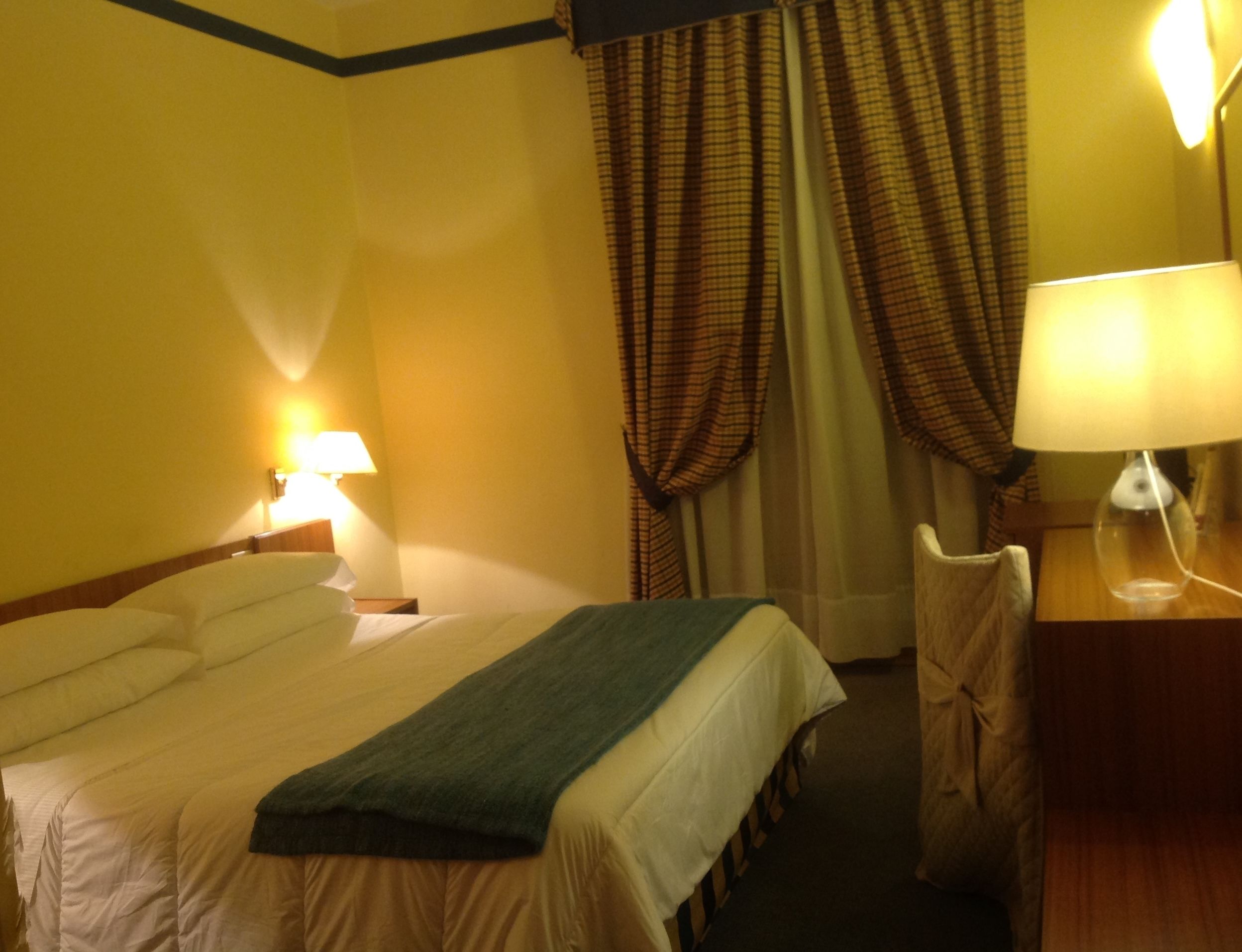 A hotel room with a bed, two bedside lamps, a desk with a lamp, and a chair.