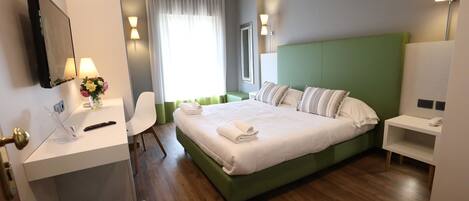 A hotel room with a green headboard, a bed with white linens, a bedside table with a lamp, a desk with a chair, and a mirror on the wall.