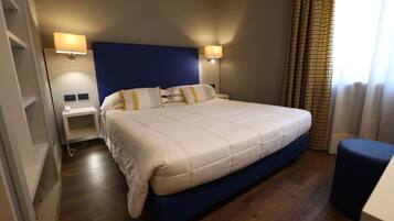 A hotel room with a large bed, two bedside tables, a nightstand, a blue ottoman, and a window with curtains.