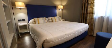 A hotel room with a large bed, two bedside tables, a nightstand, a blue ottoman, and a window with curtains.
