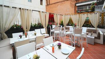 Un patio al aire libre con mobiliario de mimbre blanco, mesas preparadas para comer y una zona de estar acogedora.