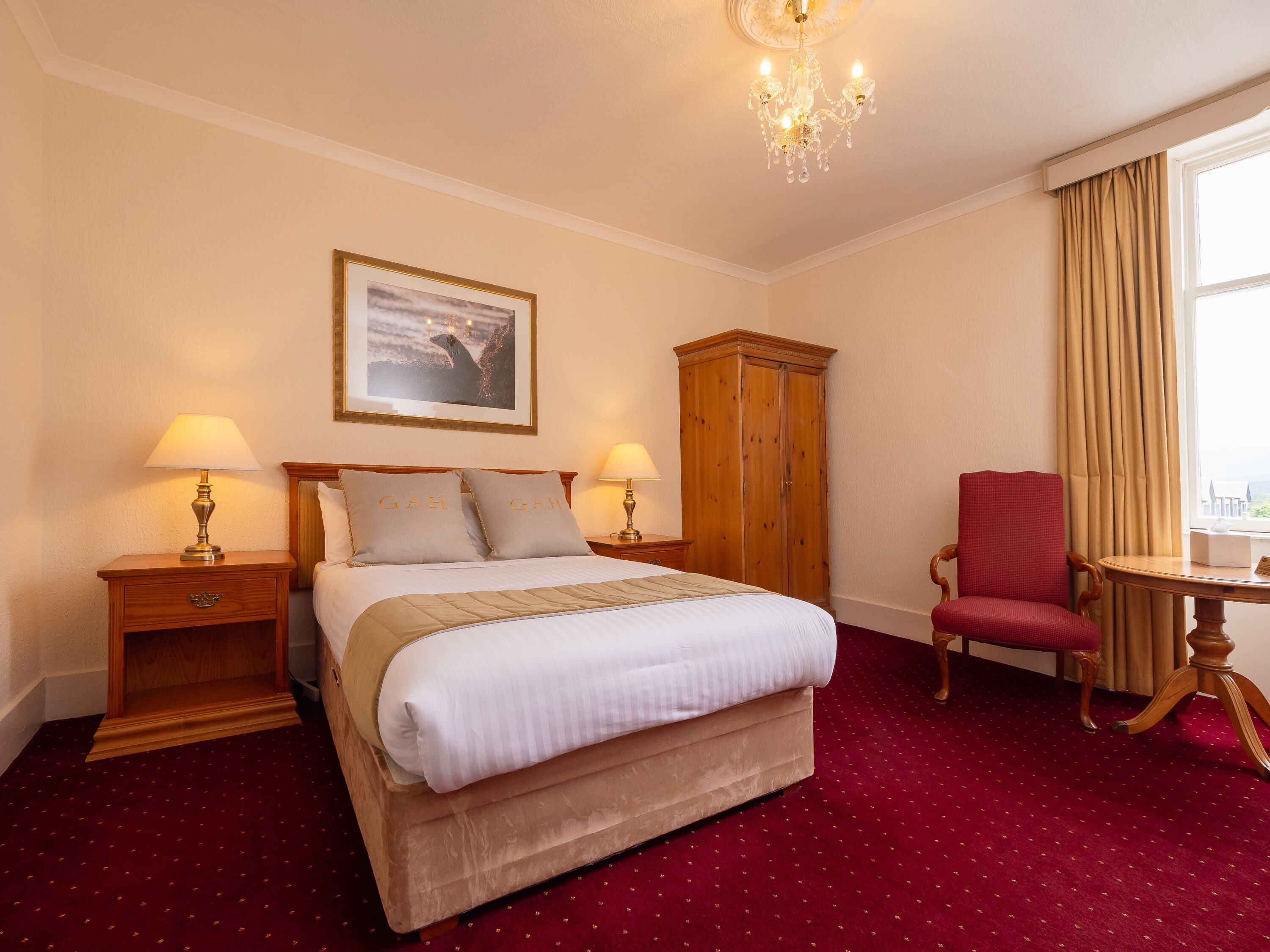 Una habitación de hotel con una cama grande, dos mesitas de noche con lámparas, un armario de madera, un sillón rojo y una ventana con cortinas.