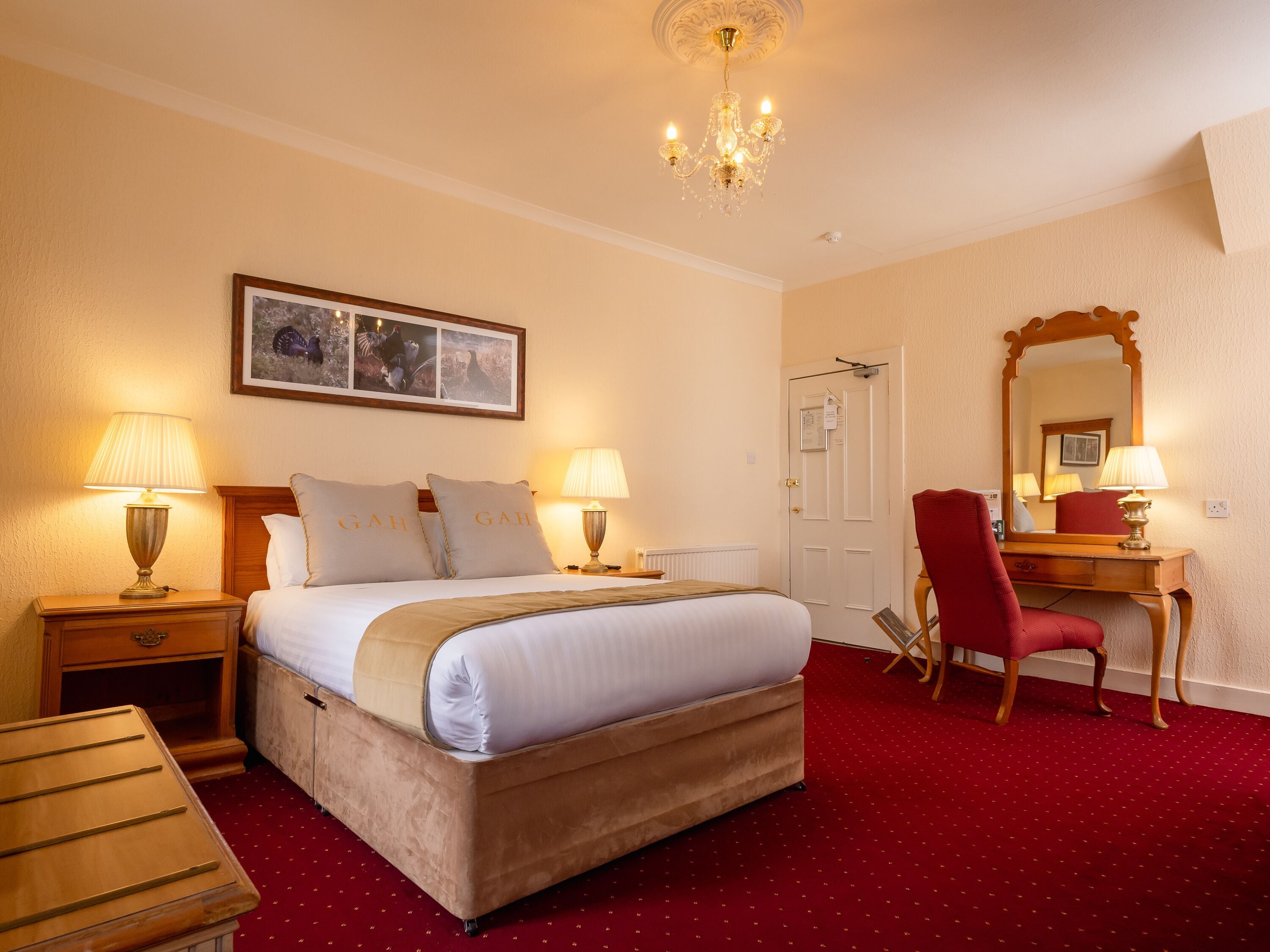 Une chambre d’hôtel avec un lit, deux lampes de chevet, une moquette rouge, un bureau en bois, une chaise rouge, un miroir et un lustre.