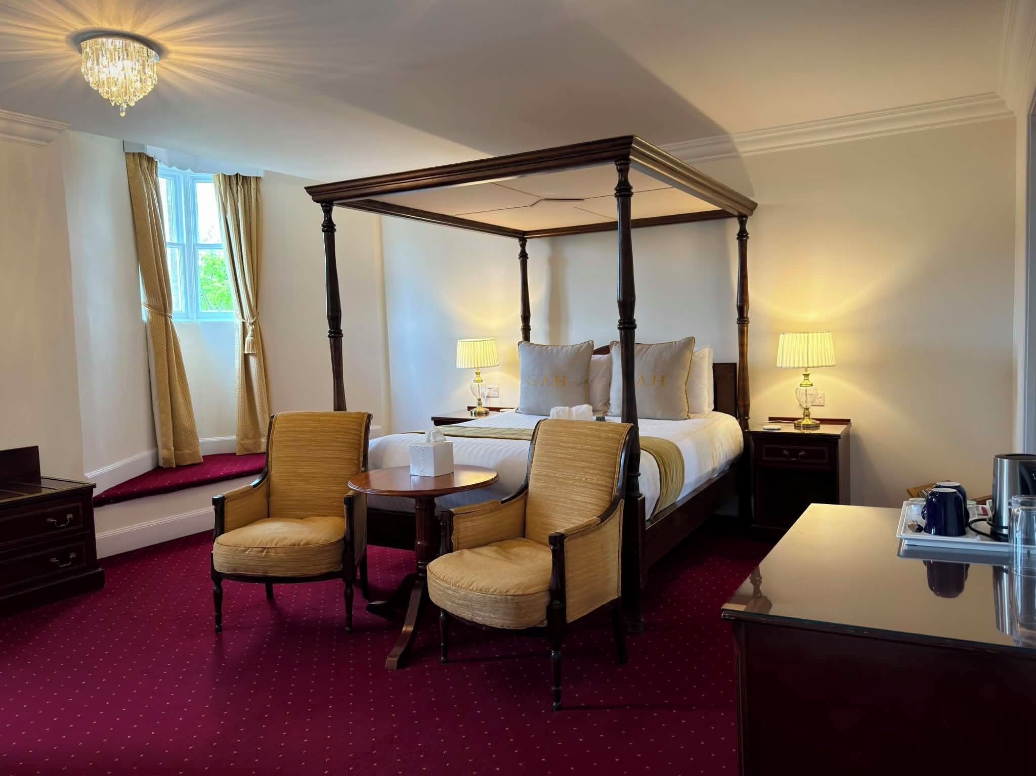 A four-poster bed with a canopy, two upholstered chairs, a bedside table with a lamp, and a small table with a cup and a kettle.