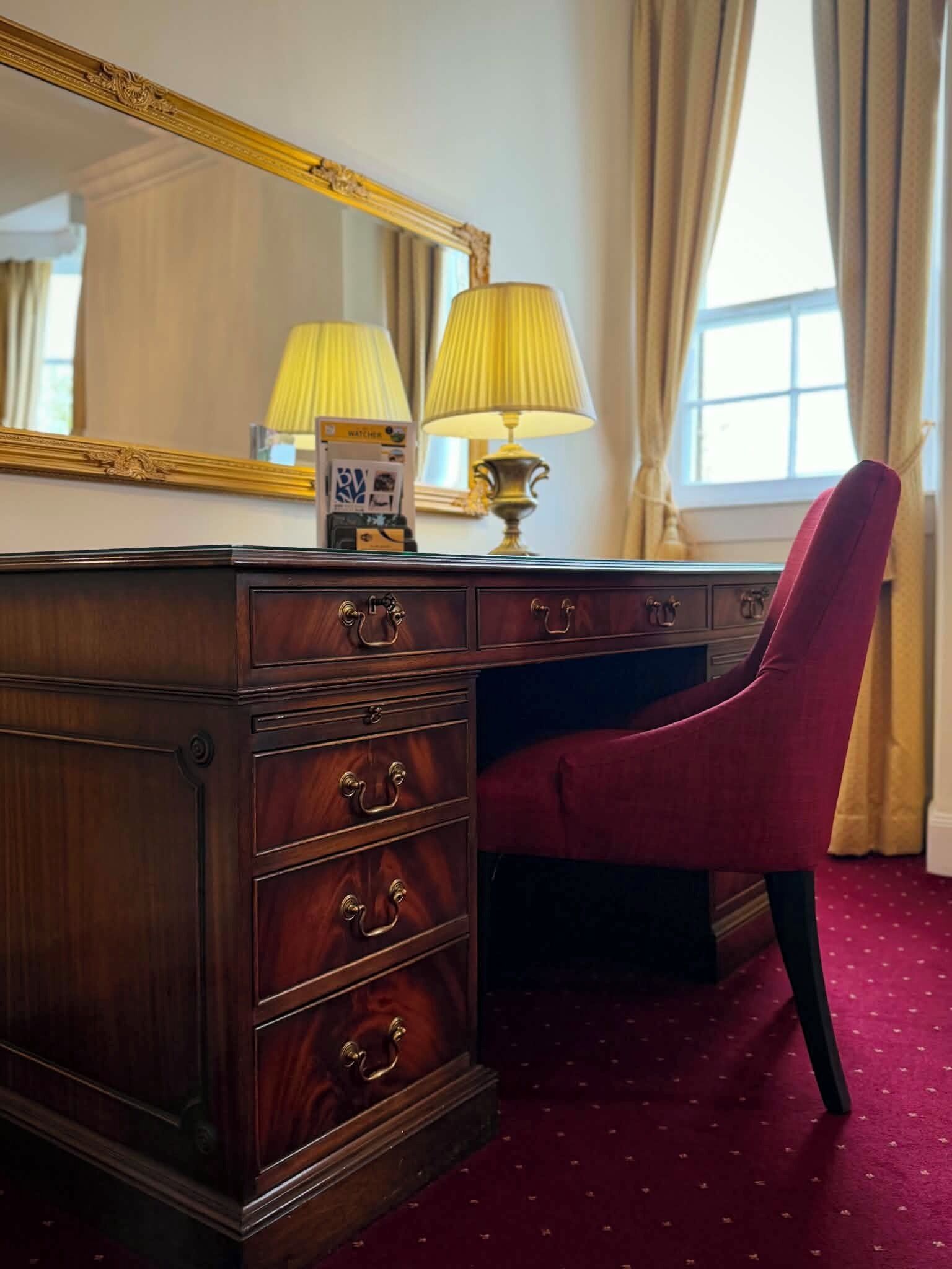 Een houten dressoir met lades en een spiegel, een rode stoel, een lamp en een raam met gordijnen.