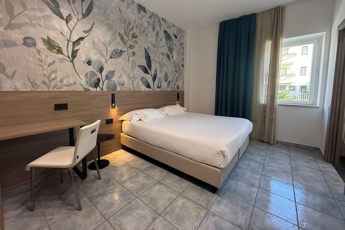 Une chambre d’hôtel comprenant un lit, un bureau, une chaise, un grand décor mural représentant des motifs floraux et une fenêtre avec des rideaux.