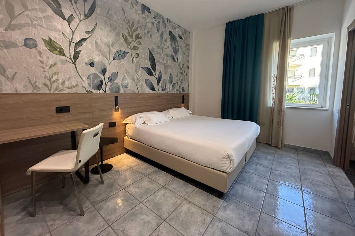 Une chambre d’hôtel avec un lit, un bureau, une chaise, un grand mur décoré de motifs floraux et une fenêtre avec des rideaux.